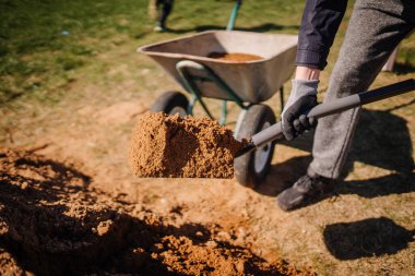 Valmiera, Letonya - 21 Nisan 2024 - Güneşli bir günde bir insan bir el arabasına toprak küreyerek bahçıvanlık veya inşaat işlerinin bir kısmını gösteriyor.