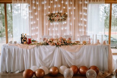 Valmiera, Letonya - 5 Ağustos 2023 - Peri ışıkları ve büyük pencereli bir odada beyaz kumaş, çiçek süsü, mum, şarap ve balonlarla süslenmiş bir düğün masası.