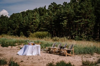 Valmiera, Letonya - 10 Ağustos 2023 - Beyaz örtülü masa, içkiler ve orman zeminine karşı ahşap sandalyeler.