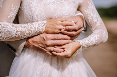 Valmiera, Latvia - August 10, 2023 - Close-up of a bride and groom's hands with wedding rings, lace dress, and suit sleeve. clipart