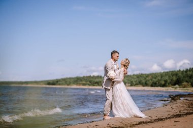 Valmiera, Letonya - 10 Ağustos 2023 - Gelin ve damat sahilde kucaklaşıyor, ayaklarının yanında dalgalar yavaşça kırılıyor, açık mavi gökyüzü.