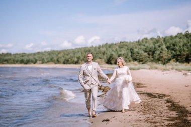 Valmiera, Letonya - 10 Ağustos 2023 - Gelin ve damat arka planda dalgalar ve ağaçlarla kumlu bir sahil boyunca el ele yürüyorlar.