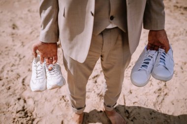 Valmiera, Letonya - 10 Ağustos 2023 - Kumlu bir plajda elinde iki beyaz spor ayakkabı tutan bir damadın yakın çekimi.