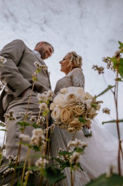 Valmiera, Letonya - 10 Ağustos 2023 - Gülümseyen gelin ve damadın alçak açılı görüntüsü, gökyüzü yukarıdayken ve gelin bir buket çiçek tutarken.