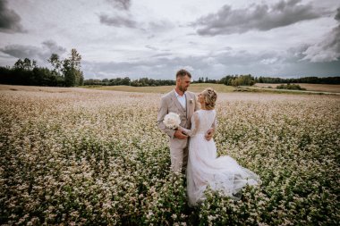 Valmiera, Letonya - 10 Ağustos 2023 Dramatik bir gökyüzünün altında beyaz çiçek tarlasında duran düğün çifti.