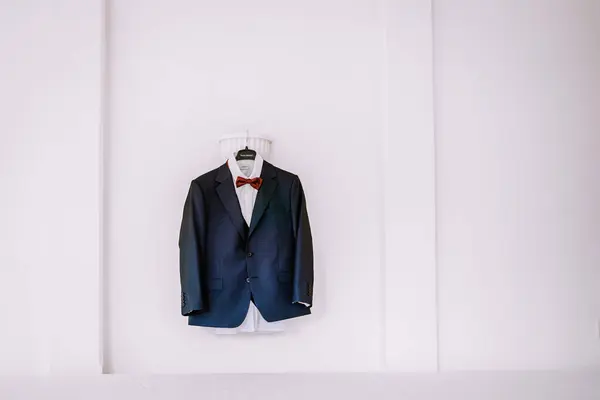 Valmiera, Latvia - August 19, 2023 - A navy blue suit with a white shirt and red bow tie, hanging on a white hanger against a clean white background.