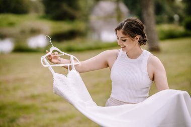 Valmiera, Letonya - 19 Ağustos 2023 - Kolsuz giysili neşeli bir kadın, açık havada sakin bir gölün kenarında tuttuğu beyaz gelinliğe hayran..