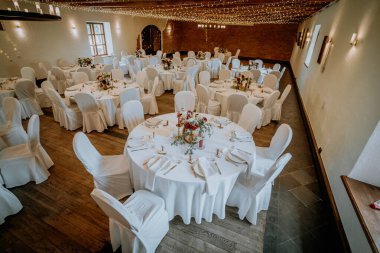 Valmiera, Letonya - 19 Ağustos 2023 - Beyaz koltuk örtüleri ve masa örtüleri olan şık bir ziyafet salonu, sicim ışıklarının altında çiçeklerle süslenmiş..