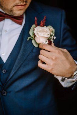 Valmiera, Letonya - 19 Ağustos 2023 - Mavi takım elbiseli bir damadın soluk gül yaka çiçeğini düzelterek kıyafetinin detaylarına odaklanması.