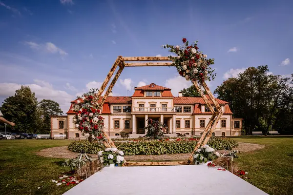 stock image Valmiera, Latvia - August 19, 2023 - An elegant outdoor wedding setup in front of a stately mansion, featuring a large wooden hexagonal arch adorned with abundant floral arrangements.