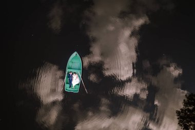 Valmiera, Letonya - 19 Ağustos 2023 - Bir gelin ve damat, sudaki ağaçlar ve bulutlarla çevrili, sakin, yansıtıcı bir su kütlesinde, yeşil bir sandalda birlikte oturuyorlar..