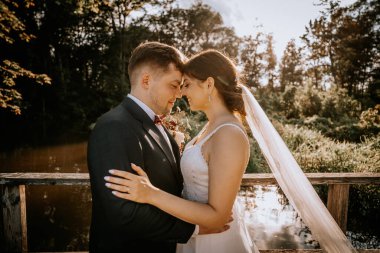 Valmiera, Letonya - 19 Ağustos 2023 - Bir gelin ve damat, arka planda yemyeşil ağaçlarla dolu bir gölün üzerindeki ahşap köprüde kucaklaşırlar..