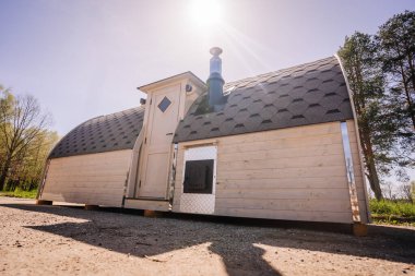 Exterior view of a modern mobile sauna with a shingled roof, a chimney, and sunlight, positioned in a natural setting. clipart