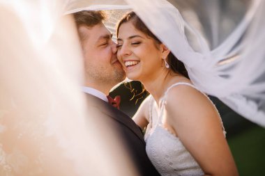 Valmiera, Letonya - 19 Ağustos 2023 - Damat gelini güneşli bir duvağın altından öpüyor, hem gülüyor, hem de düğün günlerinde samimi ve neşeli bir anda..