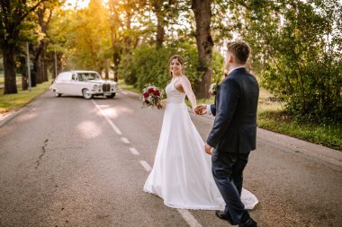 Valmiera, Letonya - 19 Ağustos 2023 - Damada gülümseyen gelin, yolda yürürken el ele tutuşan, arka planda klasik araba.