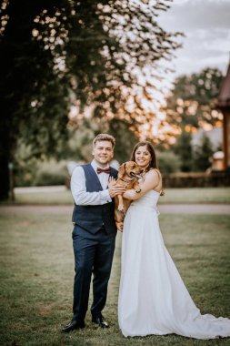 Valmiera, Letonya - 19 Ağustos 2023 - Bir gelin ve damat açık havada, kahverengi bir köpeği tutarken gülümsüyor.