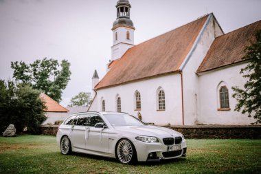 Valmiera, Letonya - 25 Ağustos 2023 - Lüks düğün arabası eski bir kilisenin önüne park edilmiş, kurdelelerle süslenmiş, yemyeşil bir arazide geçiyor..