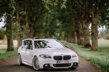 Valmiera, Letonya - 25 Ağustos 2023 Şatafatlı beyaz BMW, şık tasarım ve düğün kurdelelerini gösteren ağaçlarla kaplı manzaralı bir yolda park etmiş..