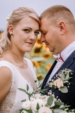 Valmiera, Letonya - 25 Ağustos 2023 - Bir gelin ve damadın yakın plan portresi Alnına hafifçe dokunuyor, bulanık bir ayçiçeği tarlası arka planına.