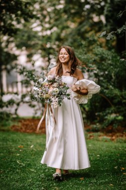Riga, Letonya, - 26 Ağustos 2024 - Gelin gülümsüyor, büyük bir buket tutuyor, yemyeşil bir parkta yürüyor.