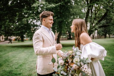 Riga, Letonya - 26 Ağustos 2024 Gelin ve damat parkta gülüyor, damat lavanta tutuyor.