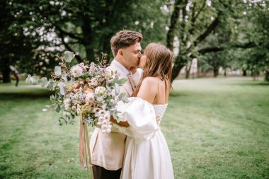 Riga, Letonya - 26 Ağustos 2024 - Gelin ve damat parkta öpüşüyor, gelin büyük bir buket çiçek tutuyor.