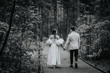 Riga, Letonya, - 26 Ağustos 2024 - Gelin ve damat, yemyeşil bir ormanda ahşap bir yolda el ele yürüyorlar..