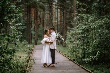 Riga, Letonya, - 26 Ağustos 2024 - Gelin ve damat yemyeşil bir ormanda tahta bir yolda kucaklaşıyorlar, gelin elinde buketle.