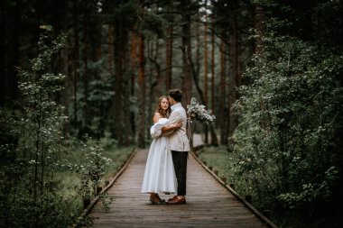 Riga, Letonya, - 26 Ağustos 2024 - Gelin ve damat yemyeşil bir ormanda tahta bir yolda kucaklaşıyorlar, gelin elinde buketle.