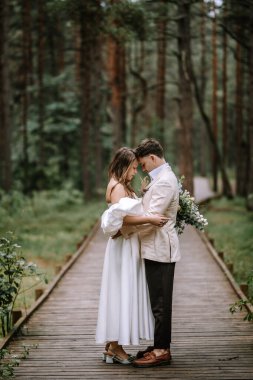 Riga, Letonya, - 26 Ağustos 2024 - Gelin ve damat yemyeşil bir ormanda tahta bir yolda kucaklaşıyorlar, gelin elinde buketle.