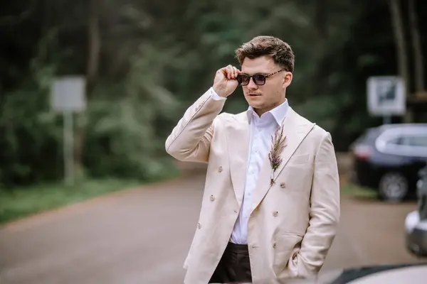 stock image Riga, Latvia, - August 26, 2024 - Stylish man adjusting sunglasses outdoors, wearing a beige suit with a floral boutonniere.