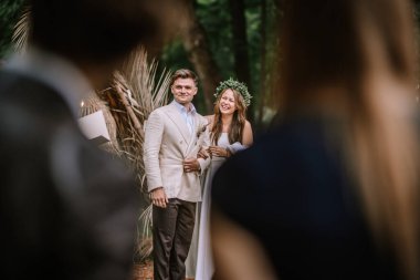 Riga, Latvia, - August 26, 2024 - A bride and groom share a joyful moment during their wedding ceremony in a lush outdoor setting, witnessed by friends and family.