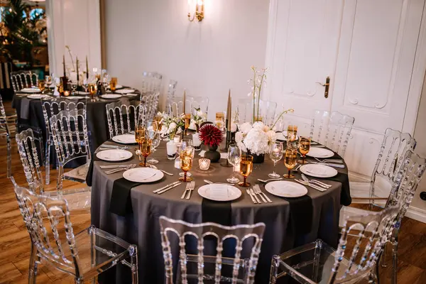 stock image Riga, Latvia, - August 26, 2024 - Sophisticated wedding reception setting featuring black tablecloths, gold-rimmed glassware, floral centerpieces, and clear chairs in an elegant room.