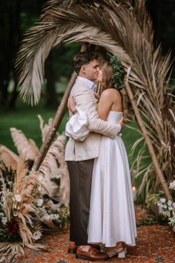 Riga, Letonya, - 26 Ağustos 2023 - Bir gelin ve damat, dekoratif bitkiler ve yeşilliklerle çevrili açık hava düğün törenlerinde öpüşürler..