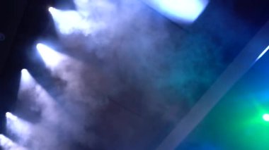 Mavi ve yeşil sahne ışıkları dumanı aydınlatarak dramatik ve atmosferik bir sahne yaratıyor..