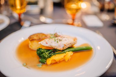 Balık filetosu, kızarmış patates, brokoli ve soslu gurme tabağı, yemek düzeninin bulanık arka planına sahip beyaz bir tabakta zarif bir şekilde servis ediliyor..