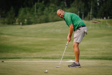 Valmiera, Letonya - 1 Haziran 2024 - Güneşli bir günde arka planda ağaçlar ve yeşillikler olan yeşil tişörtlü bir adam golf sahası kuruyor. Bulanık arkaplan.