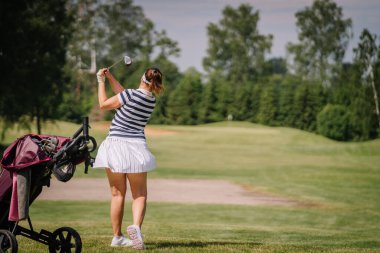 Valmiera, Letonya - 1 Haziran 2024 - Güneşli bir günde golf sopası sallayan çizgili tişörtlü ve beyaz etekli bir kadın ve arka planda ağaçlar. Bulanık arkaplan.