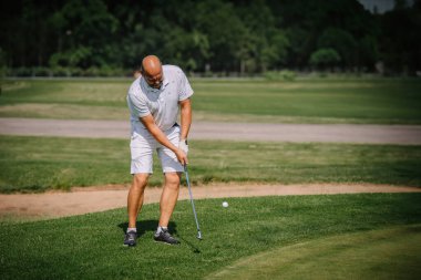 Valmiera, Letonya - 3 Haziran 2024 - Beyaz polo tişörtlü ve şortlu bir adam golf sahasında golf sopasını sallıyor, güneşli bir güne odaklanıyor.