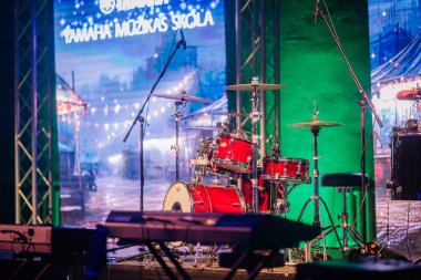 Valmiera, Latvia - May 31, 2024 - A red drum set on a stage with a colorful backdrop and lighting, ready for a live performance, with keyboards and microphones around. clipart