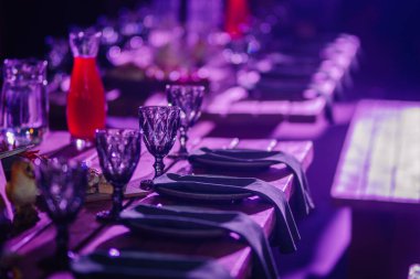 Dobele, Letonya - 7 Haziran 2024 - Mor ışık altında zarif bir yemek masası, cam eşyalar, peçeteler ve kırmızı bir içecek sürahisi, neşeli bir atmosfer yaratıyor. Boşluğu kopyala.