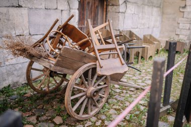 Dobele, Letonya - 7 Haziran 2024 - Kırsal sandalyeler ve kurumuş bitkilerle dolu eski ahşap bir araba, kaldırım taşlı zemini olan taştan bir duvara dayanıyor. Boşluğu kopyala.