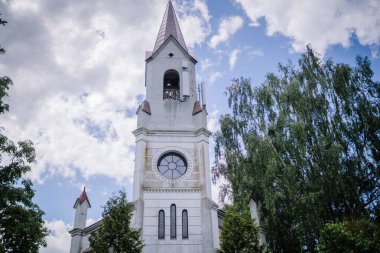 Dobele, Letonya - 7 Haziran 2024 - 5G antenli beyaz bir kilise kulesi, bulutlu ve yakındaki bir ağaçla mavi gökyüzüne kurulmuş.
