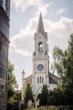 Dobele, Letonya - 7 Haziran 2024 - Uzun bir çan kulesine sahip beyaz bir kilise, bulutlarla çevrili, ağaçlarla çevrili ve ön planda bir mezarlık.