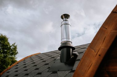 Kapalı gökyüzü olan kiremitli bir çatıdaki modern metal bacaya yakın çekim. Baca parlak ve yansıtıcıdır..