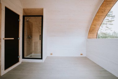 Minimalist ahşap iç döşeme, küçük bir duş odası, beyaz duvarlar ve geniş bir kopya alanı. Parlak ve temiz, doğal ışıkla.