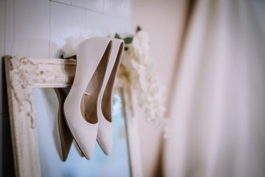 Valmiera, Letonya - 9 Eylül 2023 - Arka planda yumuşak çiçekli süslemeleri olan süslü bir ayna çerçevesine yerleştirilmiş yüksek topuklu beyaz ayakkabılar.