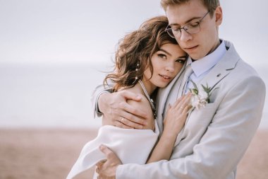 Valmiera, Letonya - 9 Eylül 2023 - Gelin ve damadın sahilde kucaklaşması, gelinin kameraya bakması ve damadın aşağı bakması, samimiyet ve aşkı vurgulaması.