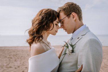 Valmiera, Letonya - 9 Eylül 2023 - Bir gelin ve damadın yakın çekimi kumlu bir sahilde samimi bir anı paylaşıyor, alınları birbirine dokunuyor ve gözleri kapalı.