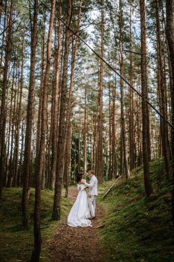 Valmiera, Letonya - 9 Eylül 2023 - Bir gelin ve damat, uzun ağaçlar ve yemyeşil ağaçlarla çevrili, sakin bir ormanda el ele tutuşup birbirlerine bakıyorlar..